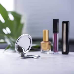 A silver pocket mirror sits open on a marble surface, with nail polish and lip products in the background.
