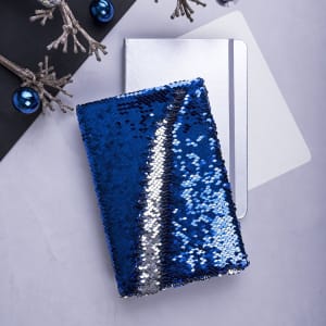 A blue and silver sequined notepad with a shiny cover, placed beside a plain white notepad and decorative items.
