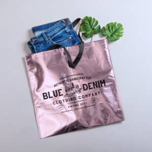 A metallic pink bag with a black handle, featuring a logo, holds a pair of blue jeans and green leaves.