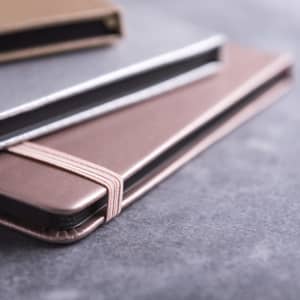 A Bodley Notepad featuring a pink cover with an elastic band, stacked with a brown notepad, on a grey background.