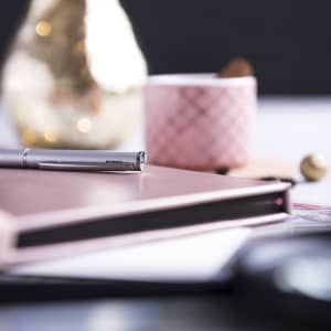 A pink notepad sits on a desk with a silver pen, a pink cup, and a decorative item in the background.