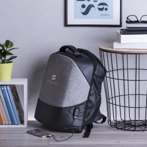 A grey and black anti-theft backpack with a logo, placed near books and a plant on a table.