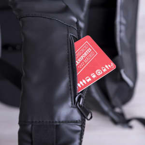 A black anti-theft backpack with a zippered pocket revealing a red card inside. It features a logo on the side.