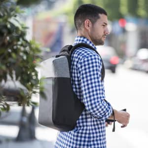 A man wearing a checked shirt carries a grey and black Ranley Anti-Theft Backpack on a city street.