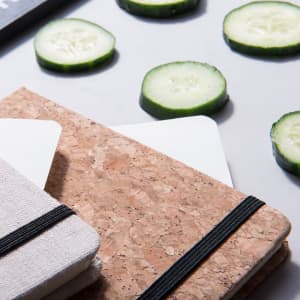 A cork notepad and a grey fabric notepad, surrounded by slices of cucumber on a light surface.