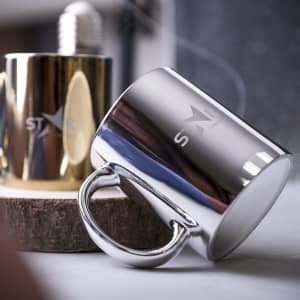 A coffee mug in shiny silver with a handle, featuring a branding logo. Another gold mug is visible in the background.