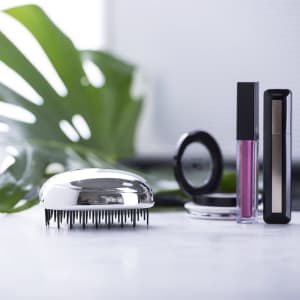 A shiny silver hairbrush alongside makeup products, including a liquid lipstick, mascara, and compact.