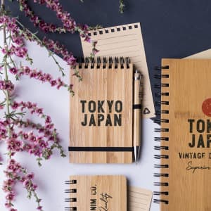 A wooden notebook with a black elastic band, accompanied by a pen and surrounded by flowers. Various notebook sizes are present.