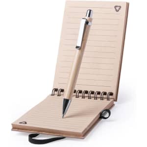 A wooden pen rests on an open, spiral-bound notebook with lined pages and a black ribbon bookmark. The notebook has a branding logo.