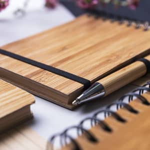 A wooden notebook with an elastic band, accompanied by a silver-inked pen, on a textured surface.