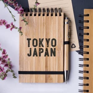 A bamboo notebook with a black elastic band and pen, accompanied by a notepad and pink flowers.