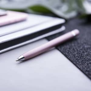 A pink stylus touch ball pen rests on a surface next to notebooks, with a blurred green plant in the background.