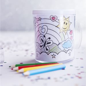 A transparent mug featuring a colouring design with rainbow, sun, hearts, and butterflies, accompanied by coloured pencils.