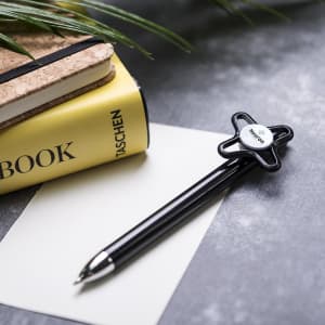 A black pen with a unique design rests on a notepad and a piece of paper, surrounded by plants.