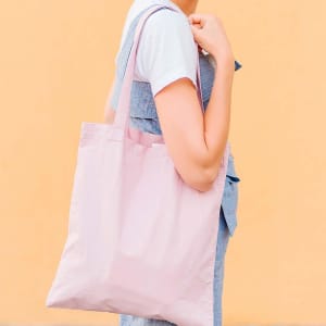 A person holds a light pink tote bag against a soft orange background. The bag features a simple design and a logo.