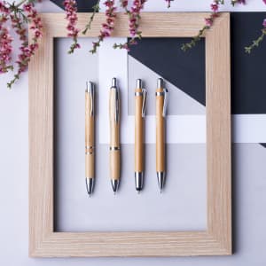 Four wooden pens with silver accents are displayed inside a light wooden frame, surrounded by pink flowers.