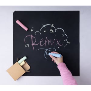 A hand writing on a black chalkboard using pastel-coloured chalks in pink, blue, and green, with a cardboard box nearby.