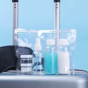 A clear toiletry bag containing spray bottles and jars in varying sizes, placed on a suitcase with a blue background.