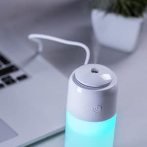 A white humidifier with a blue illuminated water reservoir, positioned next to a laptop and a small plant.