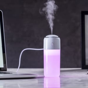 A pink and grey humidifier releasing steam, placed beside a laptop on a desk.