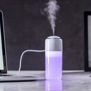 A white and purple humidifier emits mist, placed next to a laptop on a marble surface. It features a logo.