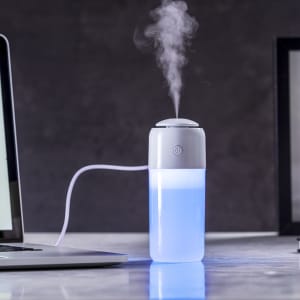 A grey and white humidifier emits steam, with a blue-lit base, placed beside a laptop on a desk.