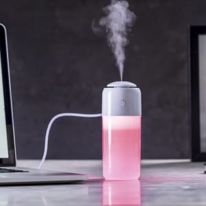 A grey and pink humidifier with mist emitting from the top, placed beside a laptop on a table.