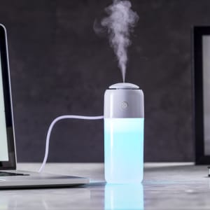 A white and light blue humidifier emitting mist, placed next to a laptop and a picture frame.