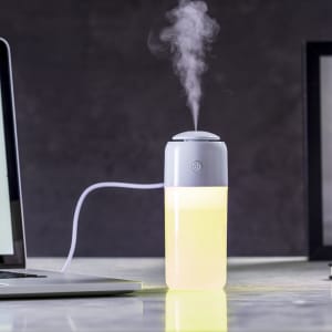 A grey and white humidifier with a glowing yellow base, emitting steam, placed on a desk near a laptop.