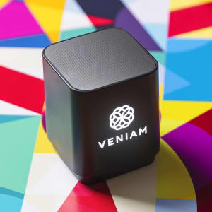 A black speaker with a square design, featuring a logo on the front, placed on a vibrant, multicoloured geometric background.
