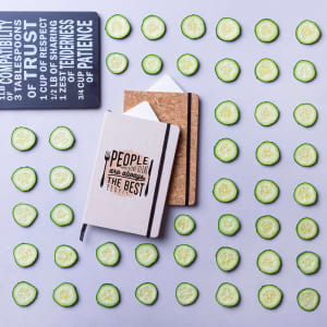 A collection of notepads in various styles, surrounded by sliced cucumbers on a flat surface. The notepads feature branding.