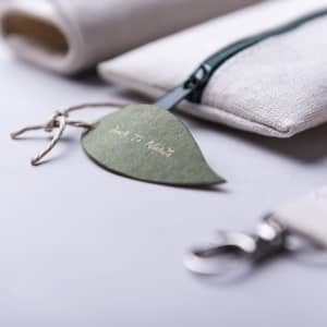 A beige pencil case with a green tag, featuring a zipper and a clip attachment. The pencil case has a simple, stylish design.