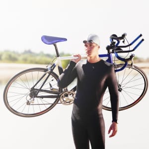 A cyclist in a wetsuit carries a bicycle on their shoulder, with a blue saddle and handlebars, against a scenic background.