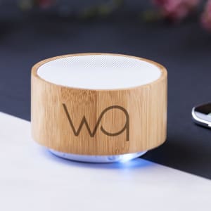A round bamboo speaker with a white mesh top, featuring a logo and illuminated base, placed on a surface.