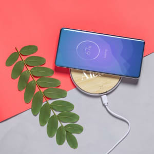A wireless charger with a wooden base and a smartphone on top, accompanied by green leaves, against a red background.