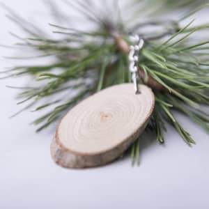 A wooden keyring with a natural finish attached to a metallic chain, set against a backdrop of green pine needles.