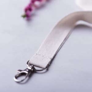 A cream-coloured lanyard with a metal clip, resting on a smooth surface with floral accents in the background.