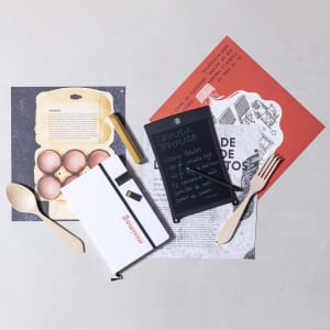 A white USB notepad with a black display, accompanied by wooden utensils and printed papers, set on a light surface.