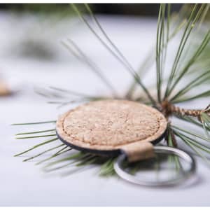 A round cork keyring with a black edge, resting on green pine needles and a light wooden surface.