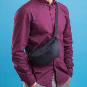 A black waistbag is worn across the body of a person in a maroon shirt, against a blue background.