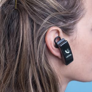 A black Bluetooth earpiece is attached to the ear of a person with long, wavy hair. It features a logo.