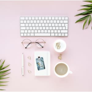 A 16Gb USB memory drive on a pink surface alongside a keyboard, glasses, a pen, a coffee cup, and greenery.