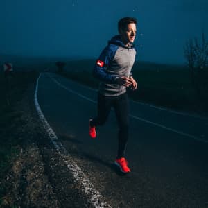 A runner in a blue hoodie and black leggings jogs along a dark road, wearing bright red shoes. A logo is visible on the hoodie.