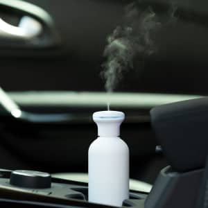 A white humidifier emits steam, placed in a car's cup holder, with a blurred background of the vehicle's interior.