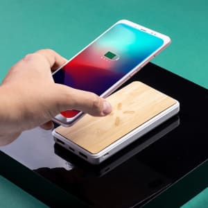 A white power bank with a bamboo top is shown, with a smartphone being placed on it against a green background.