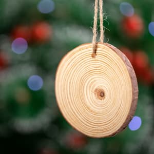 A round wooden Christmas decoration with a natural finish, hanging by a jute string, set against a blurred festive background.