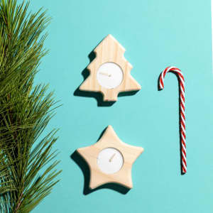 Two wooden candle holders shaped like a Christmas tree and a star, placed on a teal background with pine and a candy cane.