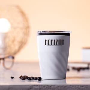 A white insulated travel mug with a black lid and a logo on the side, placed on a table with coffee beans nearby.
