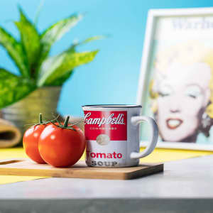 A coffee mug in red and white, featuring a logo, sits next to two tomatoes on a wooden board.