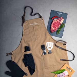 A tan apron with a logo, featuring pockets, a spatula, oven mitt, and fresh ingredients on a grey surface.